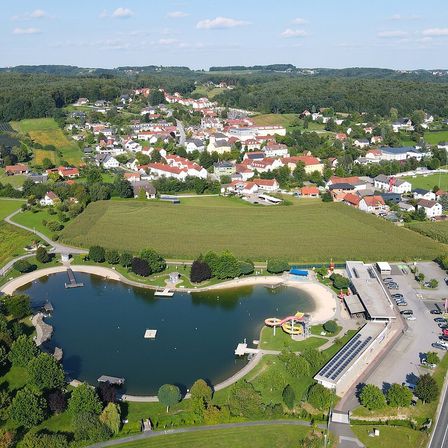 Luftaufnahme einer Stadt mit einem großen See und einem Strandbereich, umgeben von grünen Feldern und Häusern.