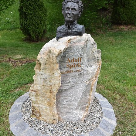 Ein Steinmonument mit einer Büste eines Mannes namens Adolf Spirk, der auf einem gespaltenen Felsen in einem Grasbereich steht, mit den Daten 1938-1993 auf dem Felsen graviert.