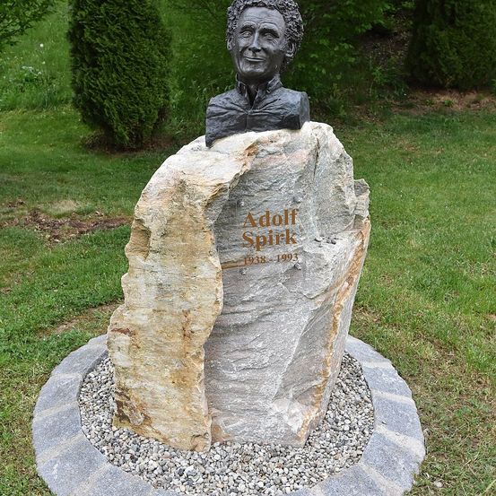 Ein Steinmonument mit einer Büste eines Mannes namens Adolf Spirk, der auf einem gespaltenen Felsen in einem Grasbereich steht, mit den Daten 1938-1993 auf dem Felsen graviert.