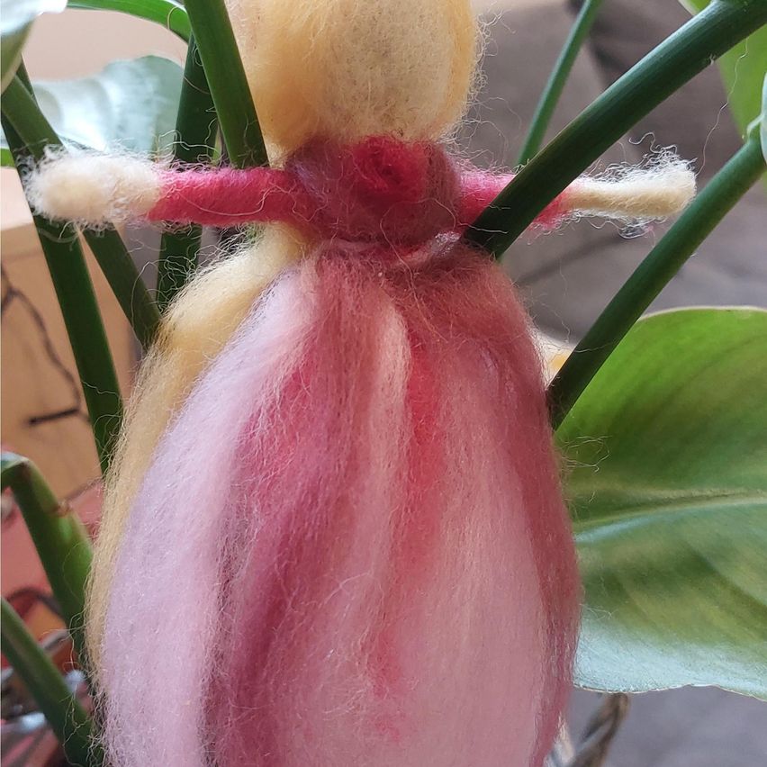 Eine kleine Puppe aus rosa und gelber Wolle sitzt auf einem Pflanzenstängel. Die Puppe hat einen rosa und weißen Körper und Arme und trägt ein rosa Kleid.