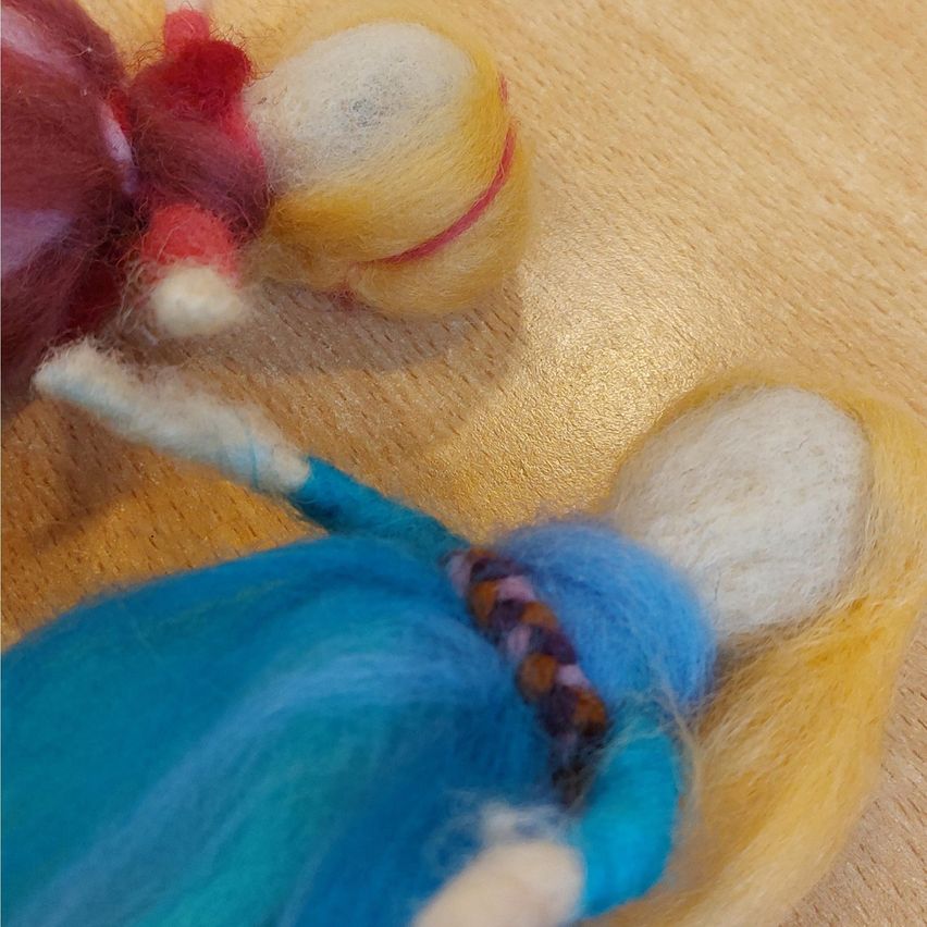 Eine Nahaufnahme von zwei Wollpuppen, eine in Blau und eine in Rot, die auf einer Holzoberfläche liegen. Die blaue Puppe hat ein geflochtenes Haaraccessoire und eine gelbe Basis.