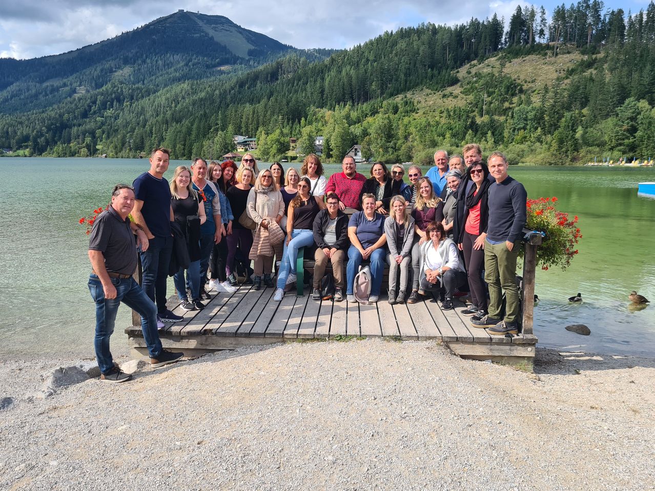 Eine Gruppe von Menschen steht und sitzt auf einem hölzernen Steg am See, mit Bergen im Hintergrund.