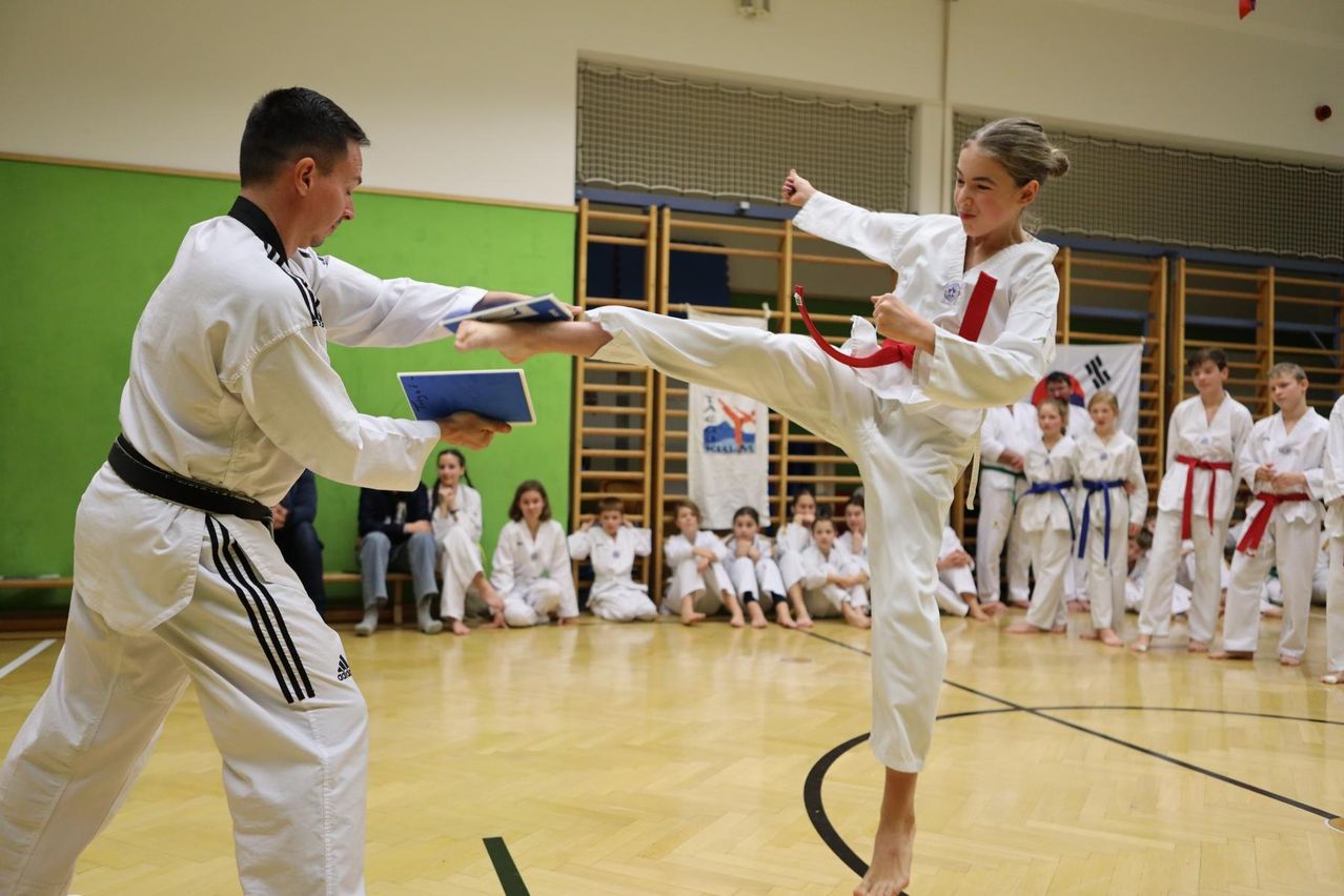 Ein Mann und ein Mädchen in Kampfsportuniformen üben das Kicken. Der Mann hält ein Buch. Sie sind in einer Turnhalle mit Kindern, die auf dem Boden sitzen.