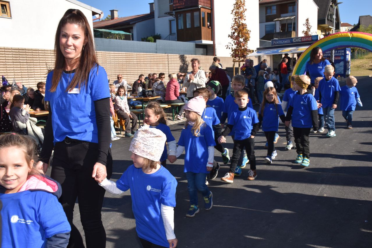 Eine Gruppe von Kindern in blauen Shirts läuft in einer Reihe, geführt von einem Erwachsenen in einem blauen Shirt. Dahinter sitzt eine Menge Menschen an Tischen und beobachtet die Kinder. Im Hintergrund sind Gebäude zu sehen.