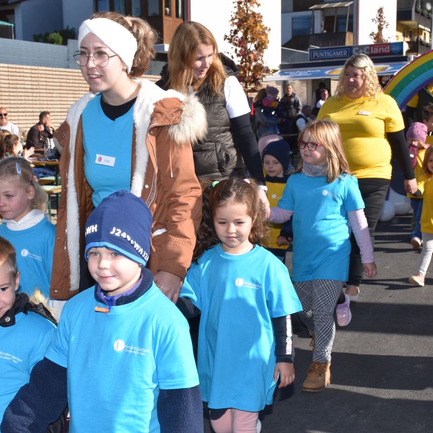 Eine Gruppe von Kindern und Erwachsenen geht auf einer Straße, trägt blaue Hemden. Einige tragen Mützen und Brillen. Dahinter sind verschwommene Menschen und Gebäude.