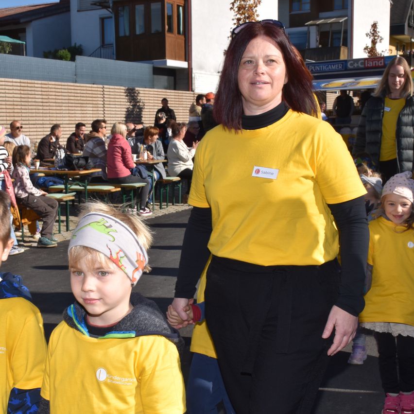 Eine Frau in einem gelben T-Shirt geht mit zwei Kindern, eines mit einem Dinosaurier-Kopfband, in einer Stadtmitte, wo Menschen an Tischen sitzen.