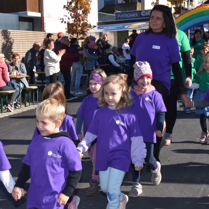 Eine Gruppe von Kindern in lila Hemden geht auf einer Straße, während Erwachsene, einschließlich einer Frau, zusehen. Im Hintergrund steht ein Gebäude mit einem Schild, das 'Puntigamer' liest.