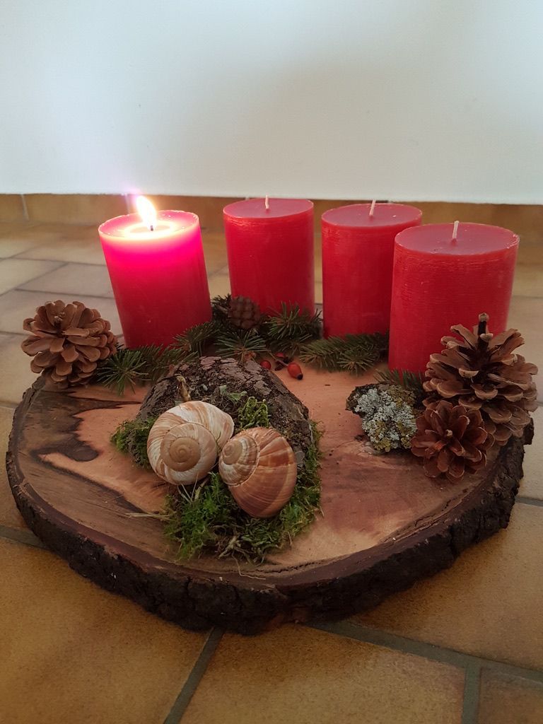 Eine hölzerne runde Basis hält vier rote Kerzen und verschiedene Kiefernzapfen und Moos, zwei Schnecken sind in der Mitte platziert.