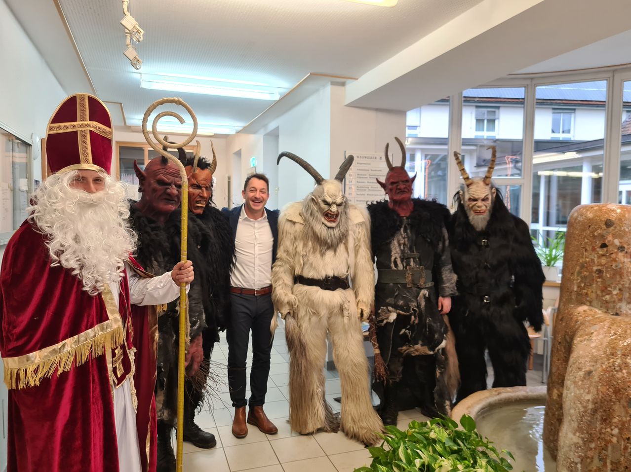 Ein Mann in einem Anzug steht mit fünf anderen in aufwendigen Kostümen, wahrscheinlich für eine Feier, in einem gut beleuchteten Raum.