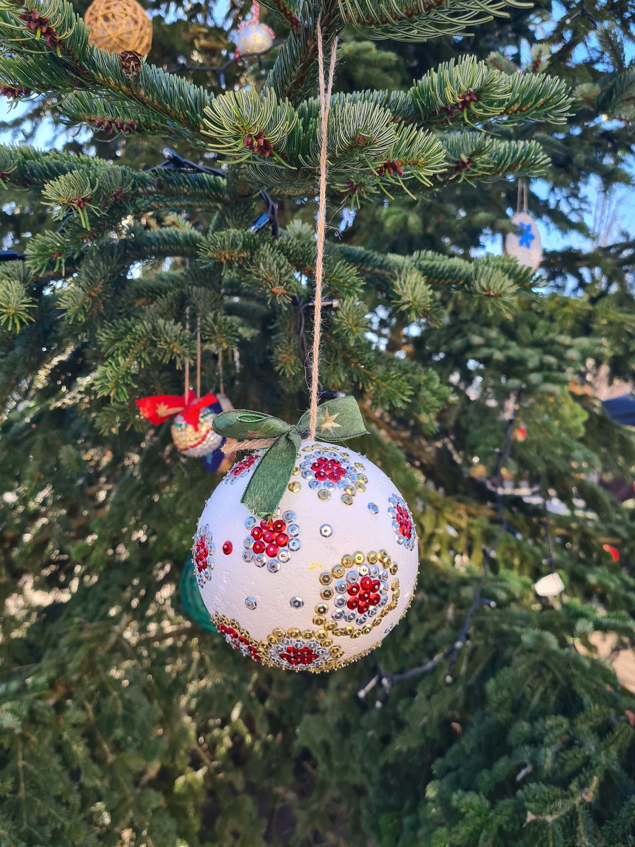 Ein weißer Weihnachtsschmuck mit roten und goldenen Mustern hängt an einem Baum. Er ist mit Pailletten und einer grünen Schleife verziert.