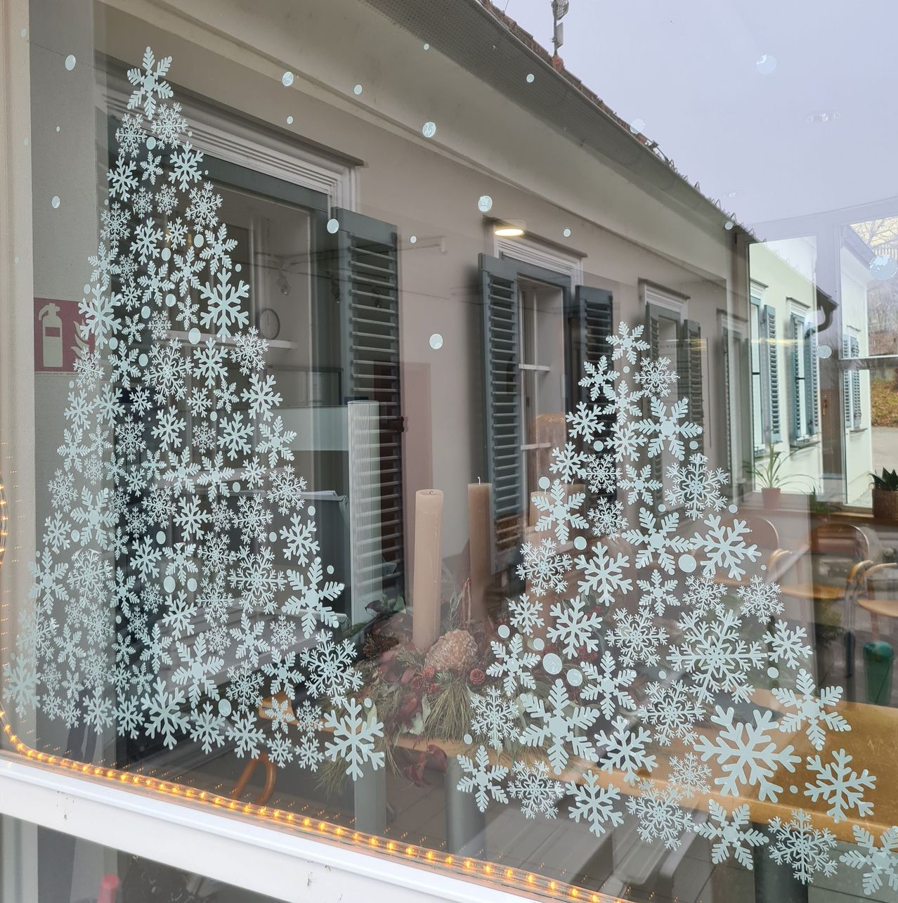 Eine festliche Szene mit zwei Weihnachtsbäumen mit Schneeflockendekorationen auf einem Fenster. Die Bäume sind mit Schneeflocken umrandet und mit gelben Lichtern beleuchtet. Es gibt einen Feuerlöscher an der Wand.