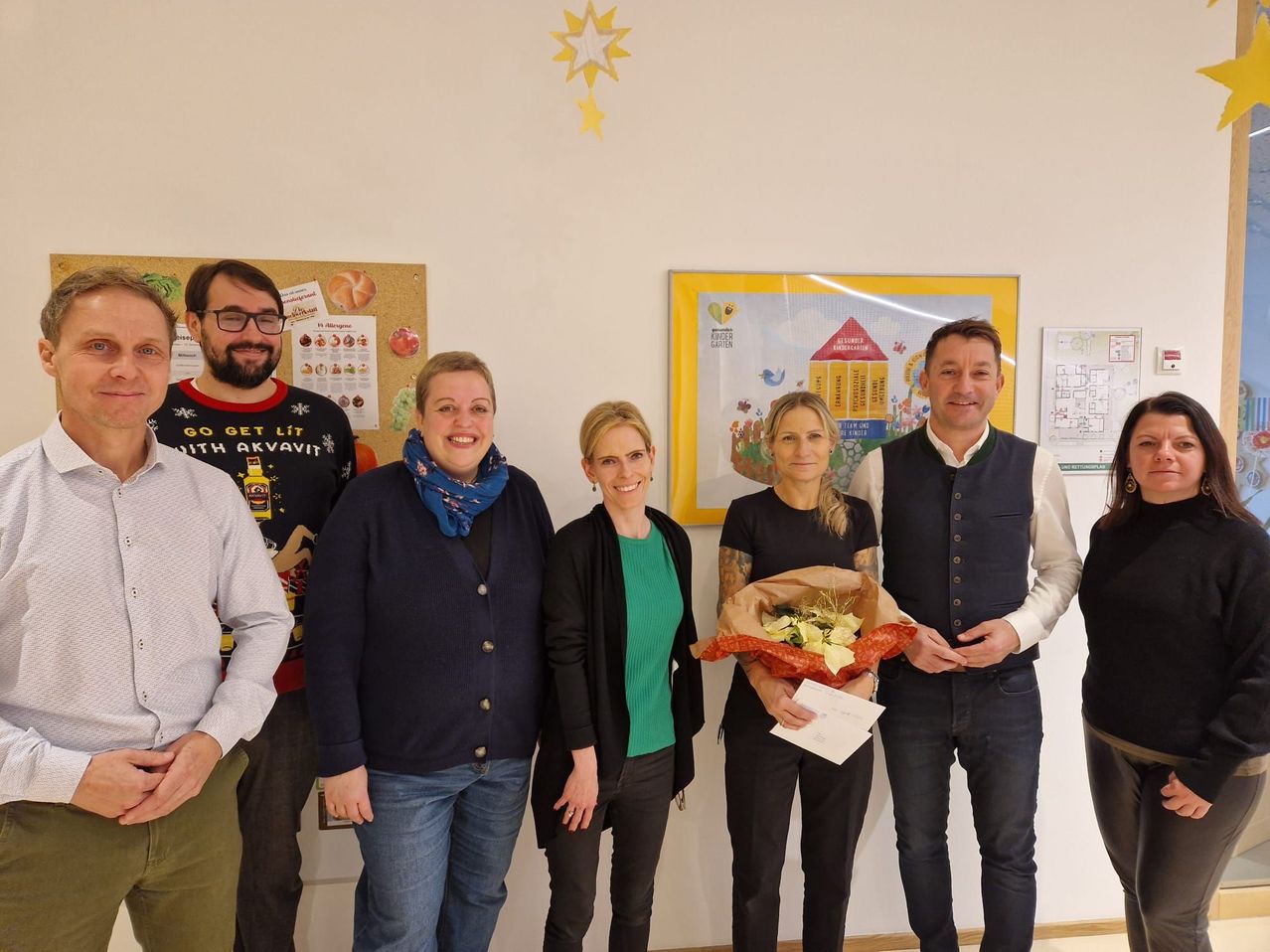 Fünf Erwachsene stehen lächelnd für ein Foto in einem Raum mit einem gelben Stern und Lehrplakaten an der Wand. Eine Frau hält einen Blumenstrauß und ein Papier.