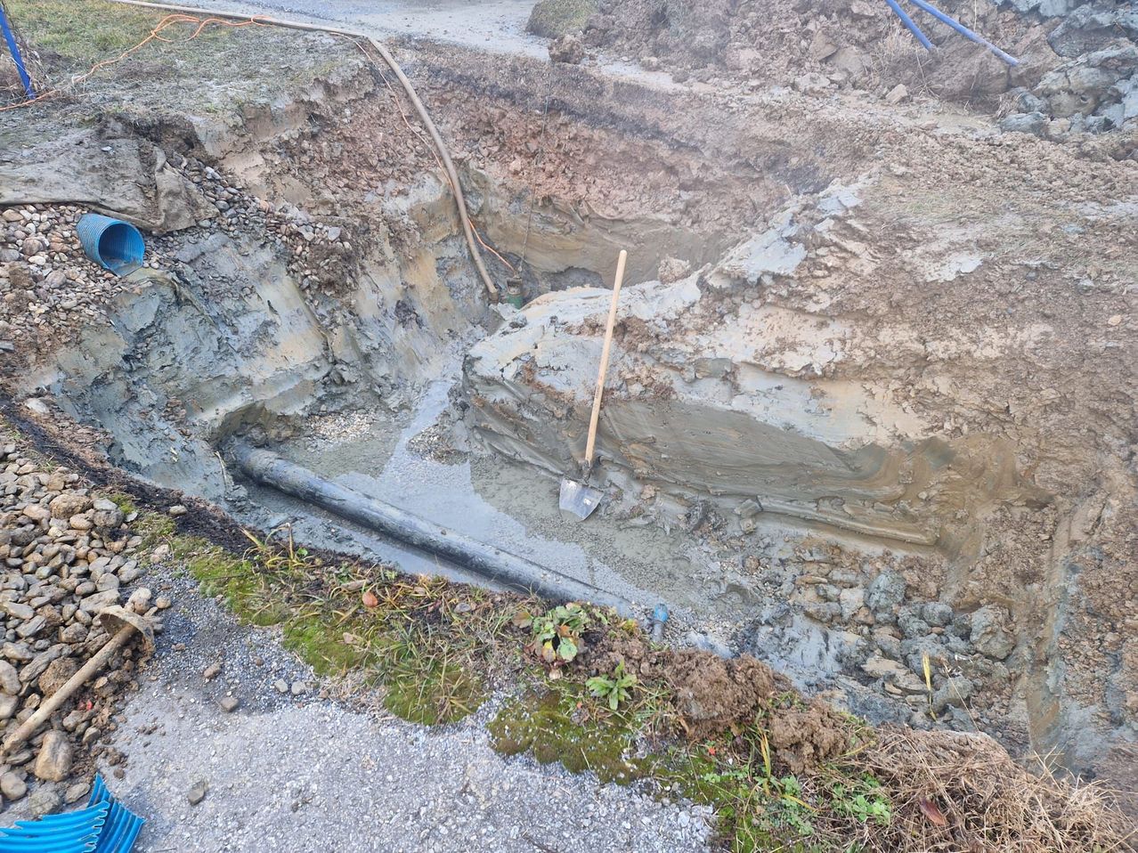 Ein Baustellenbereich zeigt eine tiefe Grube mit einem langen Rohr, das in nassem Beton eingetaucht ist. Eine Schaufel ist gegen den Rand der Grube gelehnt. Der Boden ist uneben mit freiliegendem Erdreich und verstreuten Steinen.