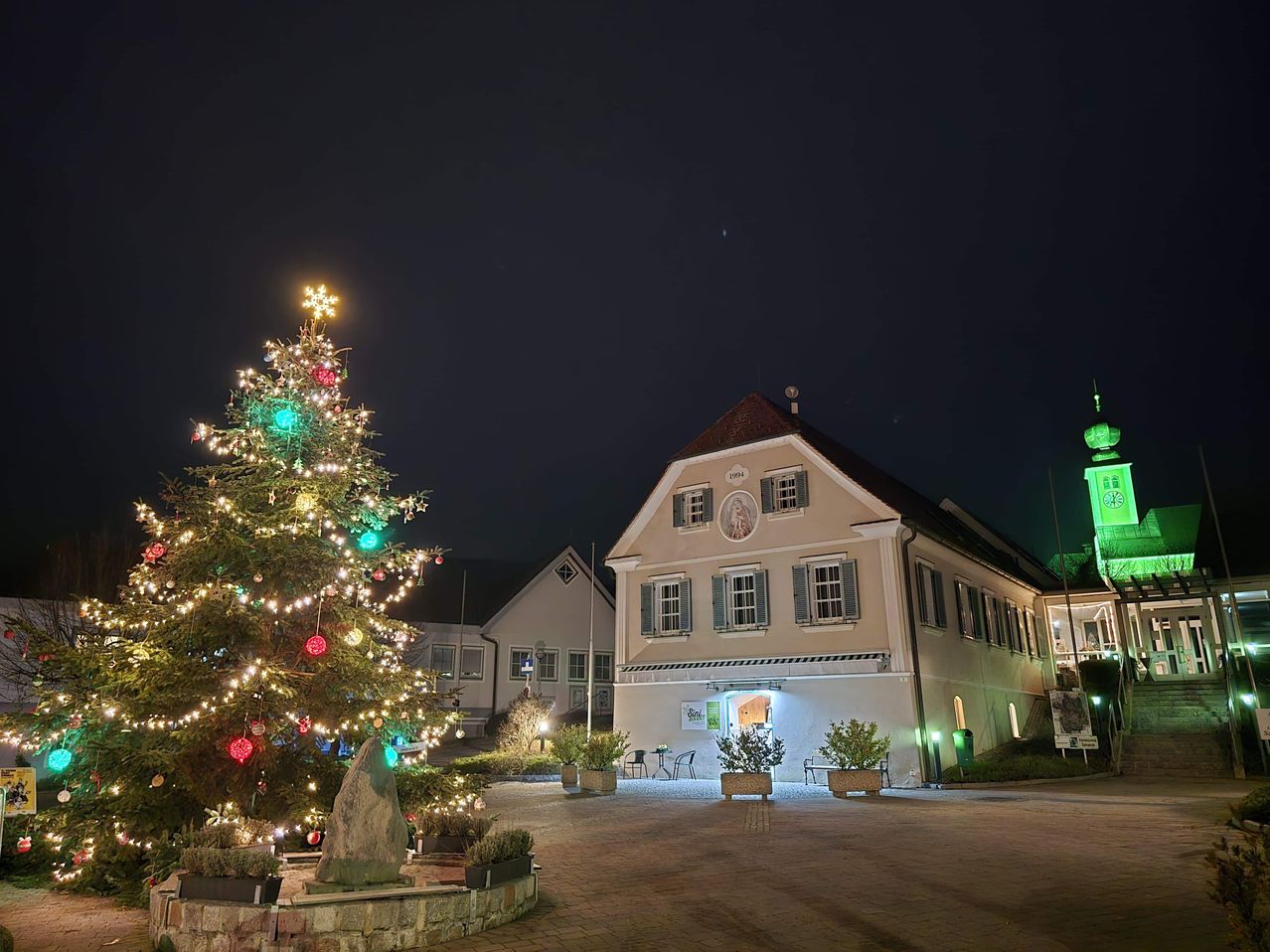 Ein Weihnachtsbaum, der mit bunten Lichtern erleuchtet ist, steht vor einem historischen Gebäude bei Nacht. Das Gebäude hat mehrere Fenster und einen grünen Turm auf der rechten Seite.