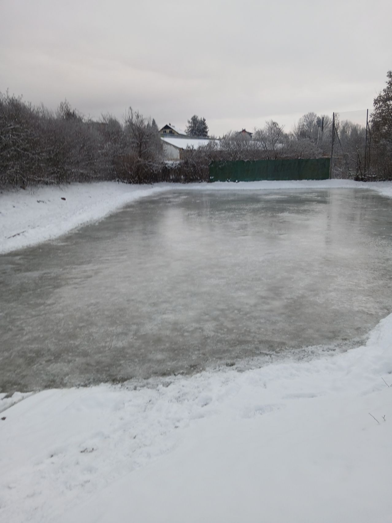 Ein zugefrorener See, umgeben von Schnee und Bäumen, mit einem grünen Zaun und entfernten Gebäuden unter einem bewölkten Himmel.