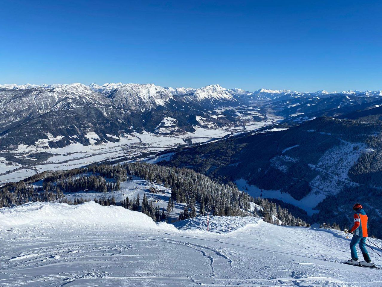 Aerial view of a snowy mountain range with trees and ski tracks. The sky is clear and blue.