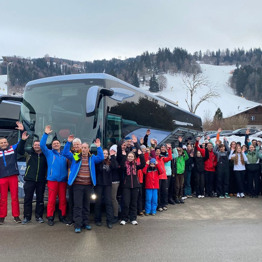 Eine Gruppe von Menschen in Winterkleidung posiert für ein Foto vor einem Bus mit einem verschneiten Berg im Hintergrund.