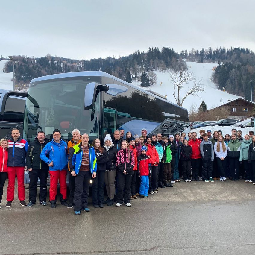 Eine Gruppe von Menschen, gekleidet in Winterkleidung, posiert für ein Foto vor einem Bus mit einem verschneiten Berg im Hintergrund.