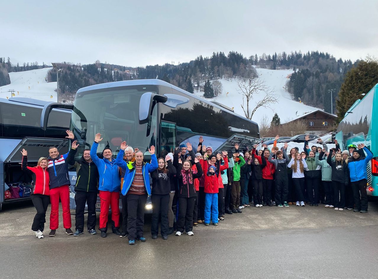 Eine Gruppe von Menschen in Winterkleidung steht vor einem Bus auf einer verschneiten Straße mit Bergen im Hintergrund.