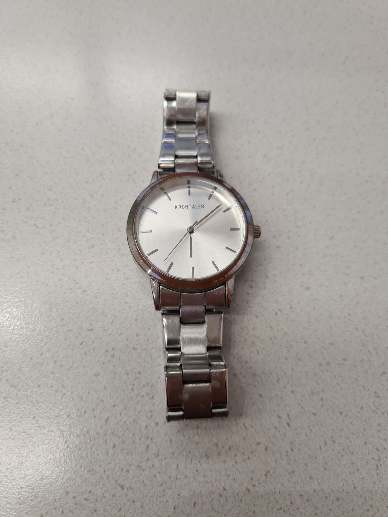 A silver wristwatch with a round face and brown strap, branded Krontaler, placed on a white surface.
