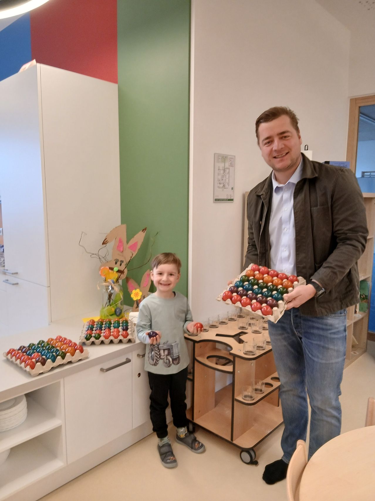 Ein Mann und ein Kind stehen in einem Raum, das Kind hält ein buntes Ei und lächelt, während der Mann ein Tablett mit bunten Eiern hält.
