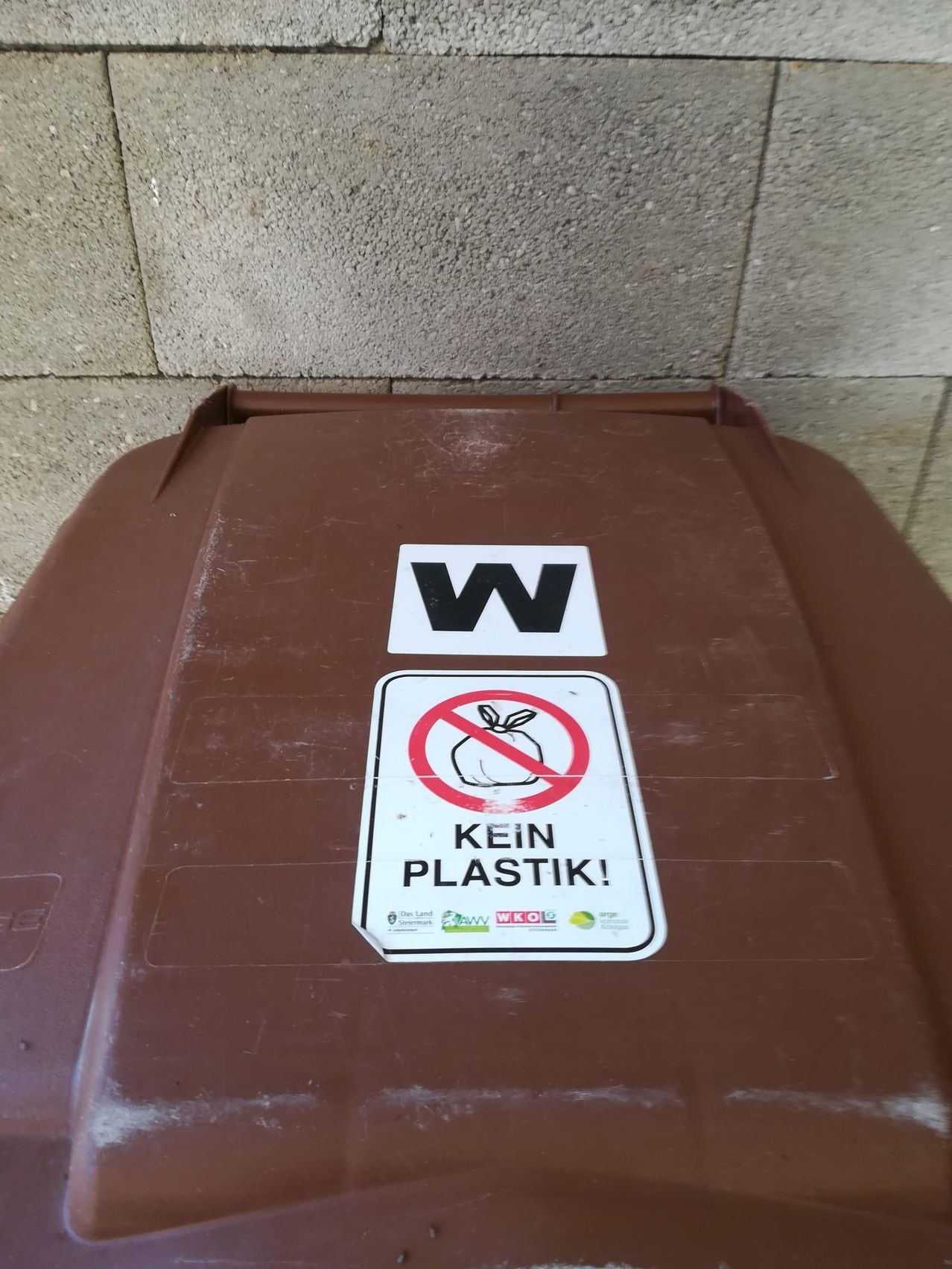 A brown waste bin with two signs. One says 'W' in black, and the other has a red circle with a slash through it and says 'KEIN PLASTIK!'.