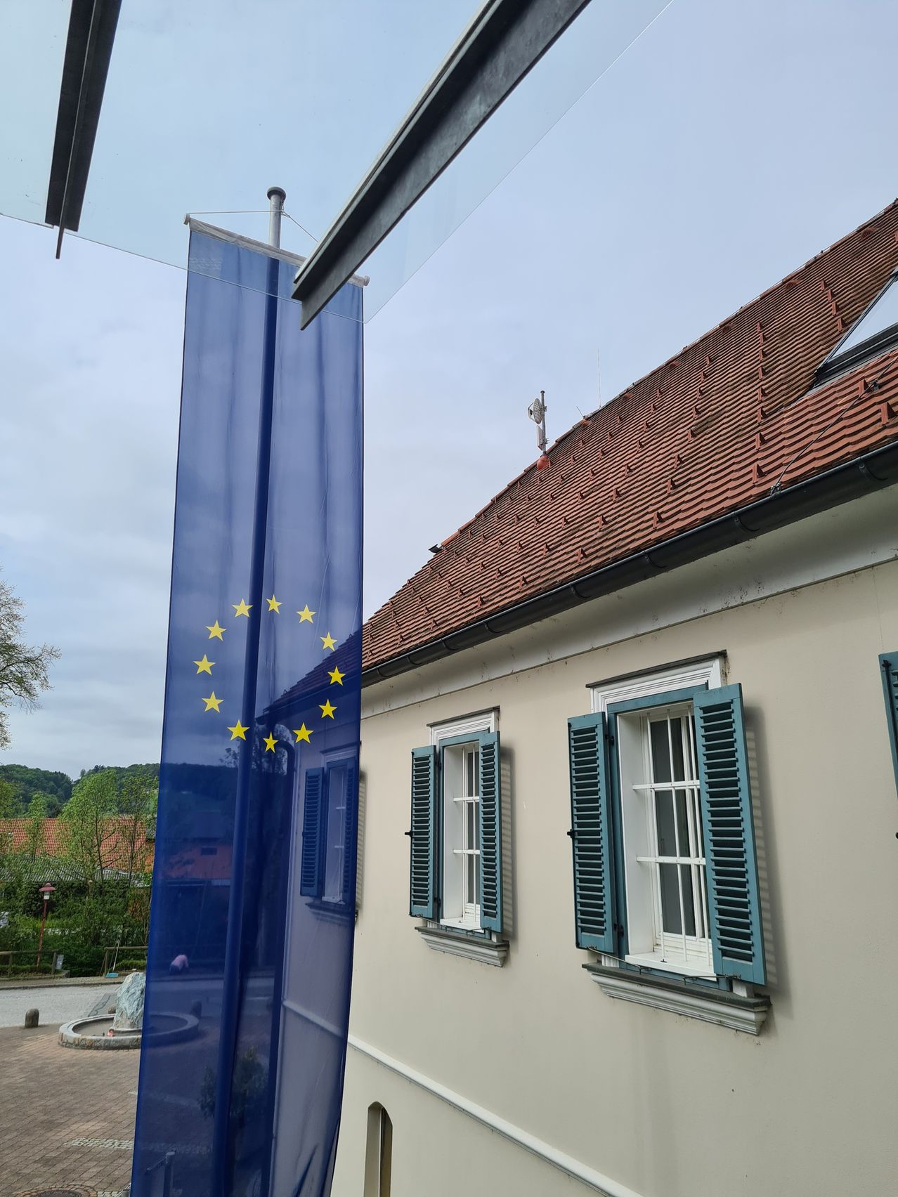 A blue European Union flag hangs next to a white building with a red roof, blue shutters, and three windows. The building is surrounded by greenery.