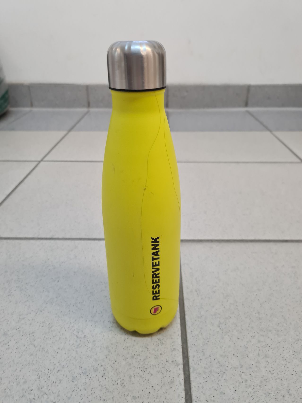 A yellow bottle with a silver lid is on a tiled floor. The bottle says 'Reservetank' on the side.