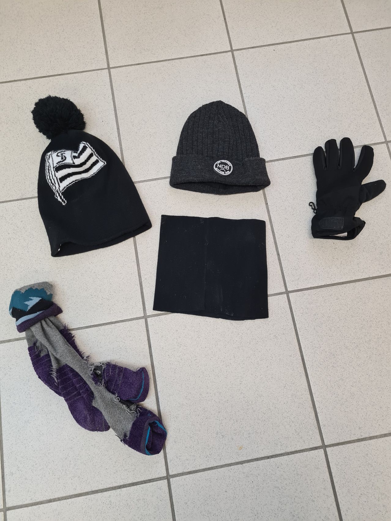 A set of winter clothing includes a black beanie with a flag, a gray beanie with an NDB logo, black gloves, a black scarf, and a pair of torn socks.