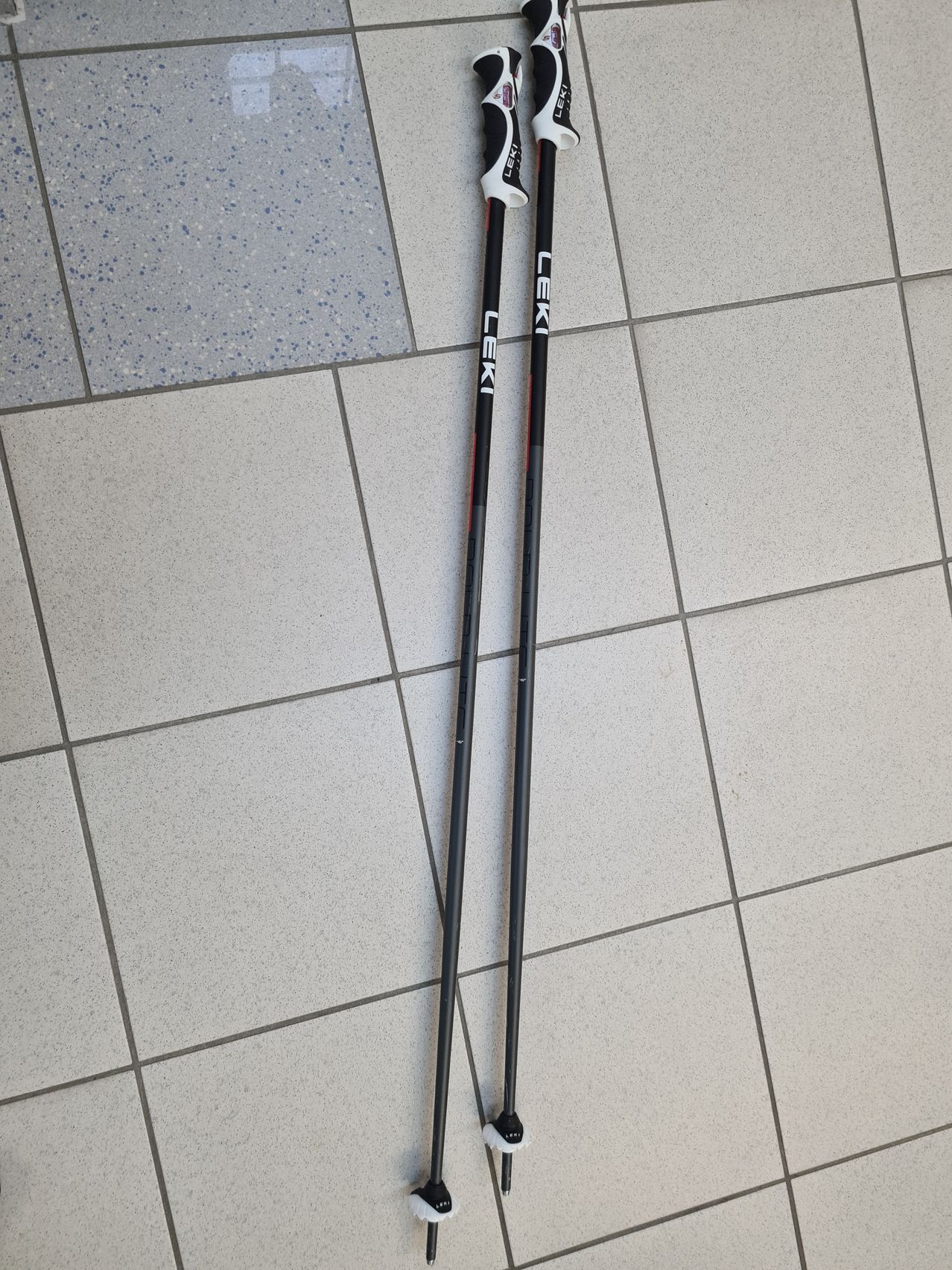 Two black ski poles are placed on a tiled floor. The poles have a white grip at the top and the word 'LEKI' written in white.