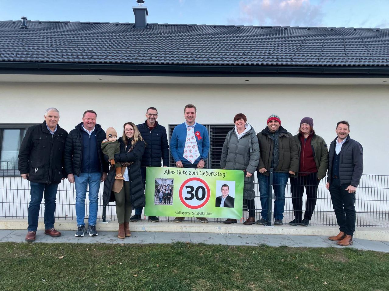 Eine Gruppe von Menschen steht vor einem Haus und lächelt für ein Foto. Sie halten ein grünes Banner mit der Nummer 30 und einem Bild eines Mannes. Das Haus hat einen Schornstein und ein Ziegeldach.