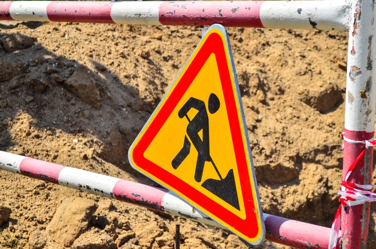 A yellow and red triangular warning sign with a silhouette of a person digging is attached to a construction barrier.