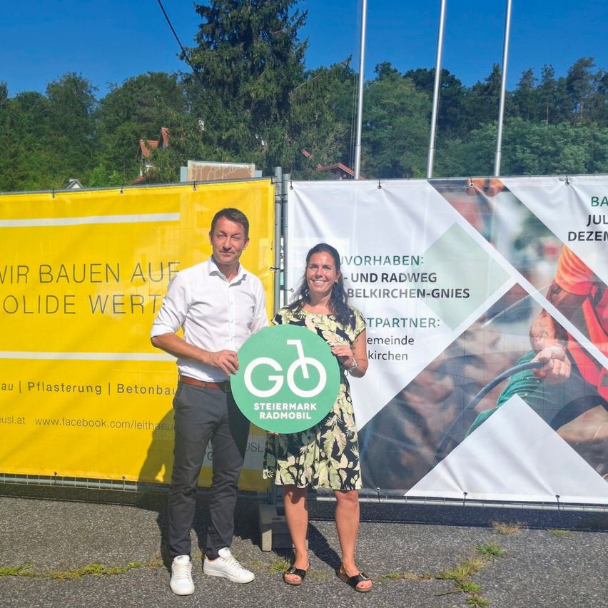 Zwei Personen stehen vor einem Banner, das eine Fahrradinitiative bewirbt, und halten ein grünes kreisförmiges Schild mit dem Text 'GD' und einem Fahrradsymbol. Der Mann trägt ein weißes Hemd und Hose, die Frau ein Blumenkleid. Dahinter steht ein Zaun mit einem gelben Banner.