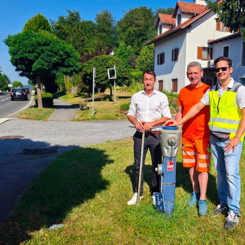 Drei Männer stehen neben einem Feuerhydranten auf einer Rasenfläche, mit einem Haus und einer Straße im Hintergrund.