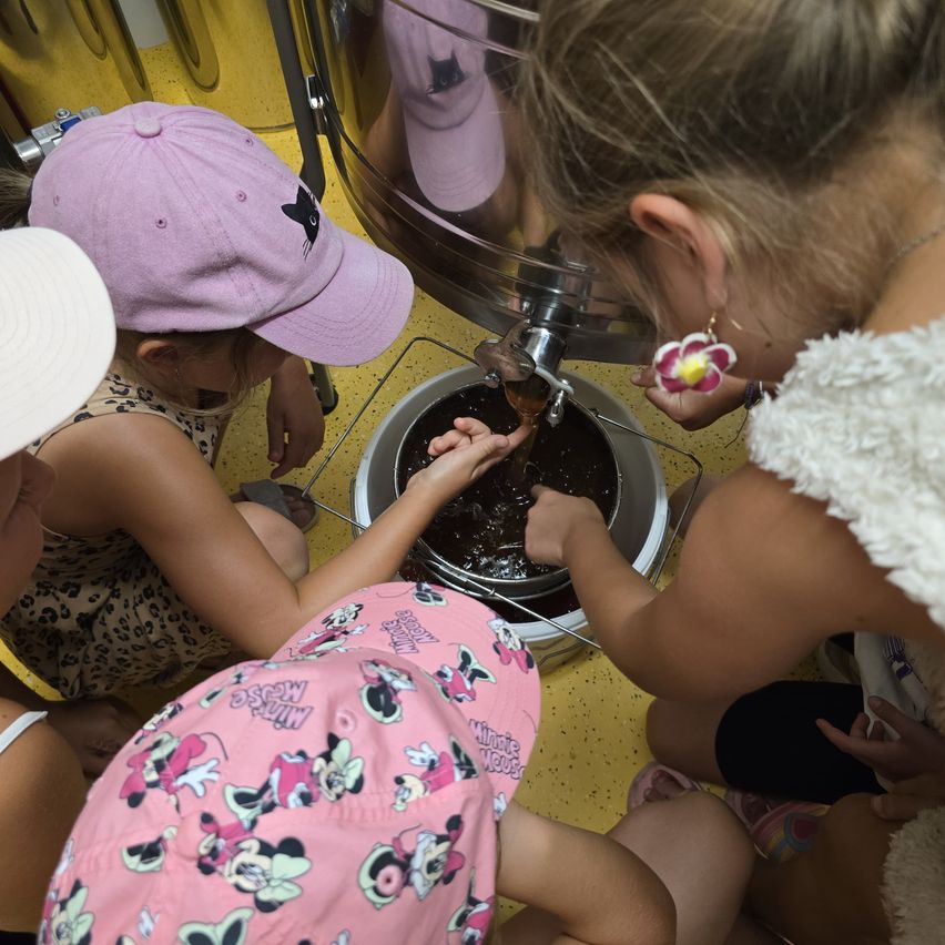 Vier junge Mädchen sind um einen großen Metallbehälter versammelt, möglicherweise in einer Schokoladenherstellungsaktivität. Jedes Mädchen trägt einen Hut, und eine gießt Schokolade in den Behälter.