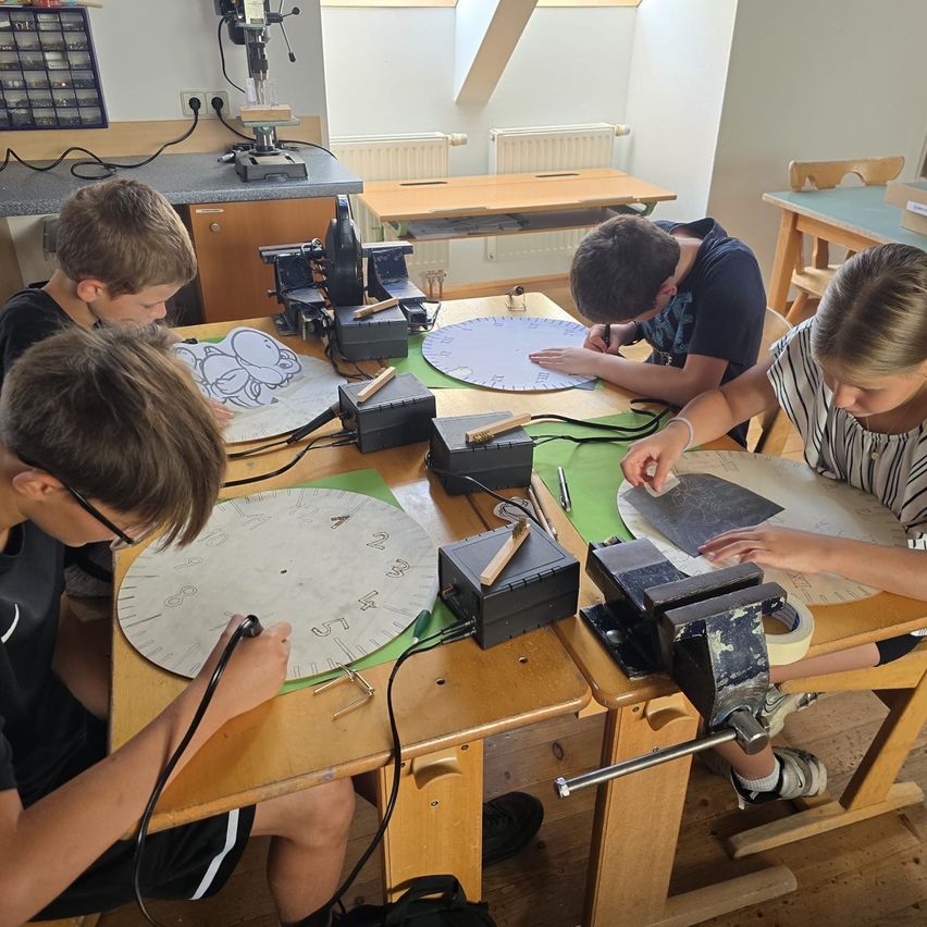 Vier Kinder arbeiten in einer Werkstatt an einem Projekt. Sie sitzen um einen Tisch herum mit verschiedenen Werkzeugen und Materialien. Jedes Kind konzentriert sich auf seine Aufgabe, wobei eines einen Stift auf einem kreisförmigen Stück Papier benutzt.