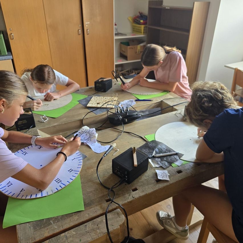 Vier Mädchen arbeiten an einem Bastelprojekt an einem Holztisch, jede mit einem kreisförmigen Papier und Zeichenwerkzeugen. Im Hintergrund befinden sich Holzschränke und Regale.