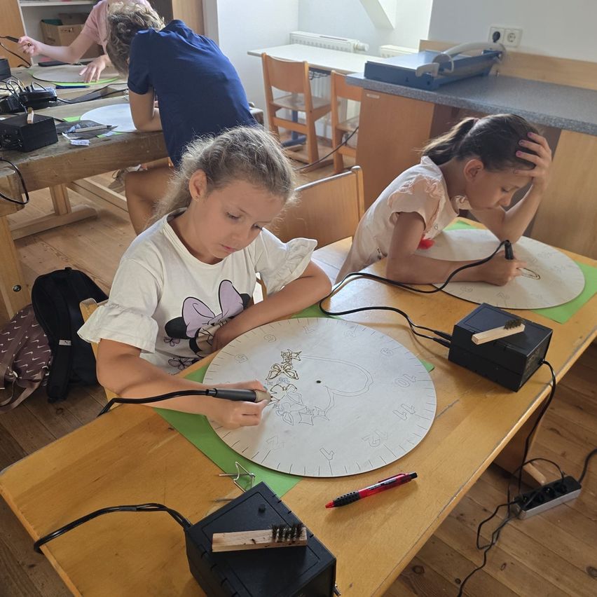 Zwei Mädchen arbeiten an einem runden Projekt an einem Tisch, eine benutzt ein Löteisen und die andere hat einen Pinsel. Sie sind von verschiedenen Werkzeugen und Materialien umgeben.
