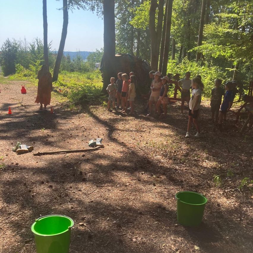 Eine Gruppe von Kindern und Erwachsenen versammelt sich in einer Waldlichtung, einige tragen Hüte. Zwei grüne Eimer stehen auf dem Boden, und orangefarbene Kegel sind verstreut. Bäume und Pflanzen umgeben das Gebiet.