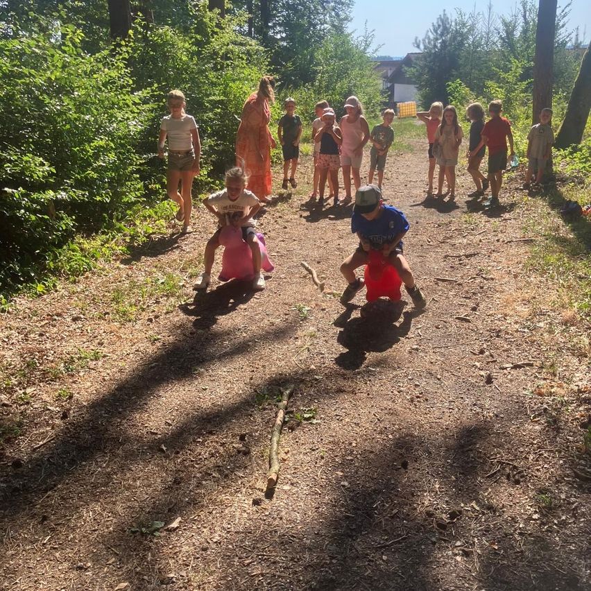 Eine Gruppe von Kindern spielt auf einem Lehmweg im Wald, einige reiten auf rosa Spielzeug.