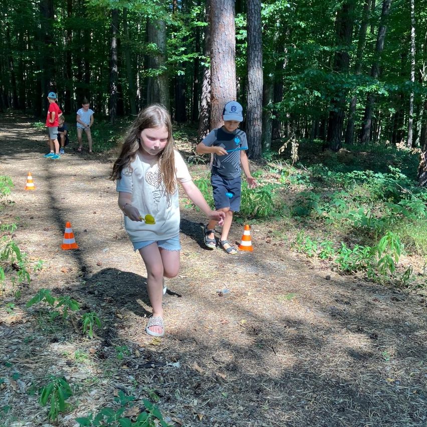 Ein Mädchen und ein Junge rennen auf einem Waldweg, wobei orangefarbene Verkehrskegel den Pfad markieren. Dahinter stehen einige Kinder und beobachten.