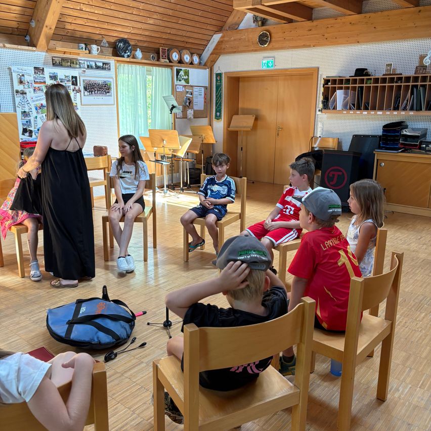 Eine Gruppe von Kindern und einer Frau sitzen im Kreis in einem Raum. Die Frau steht und scheint eine Diskussion zu leiten. Die Kinder hören aufmerksam zu. Im Hintergrund befinden sich Musikinstrumente und eine Pinnwand.
