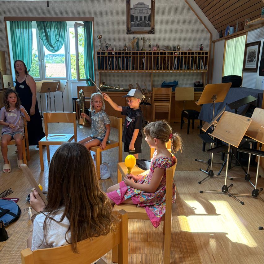 Kinder in einem Musikraum, eines hält einen Taktstock, ein anderes spielt ein gelbes Xylophon, und andere sitzen mit Instrumenten. Eine Frau steht in der Nähe eines Fensters.