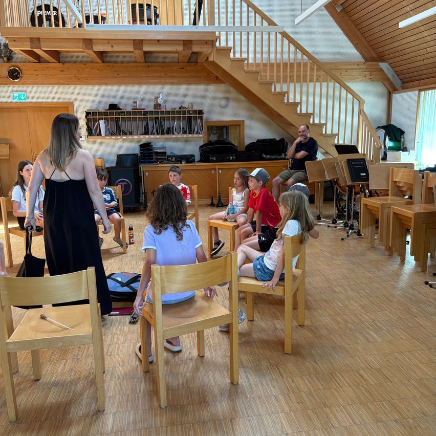 Eine Gruppe von Kindern und einer Frau sitzen in Stühlen in einem Raum mit Holzböden. Die Frau steht und scheint zu unterrichten. Musikinstrumente stehen auf dem Tisch.
