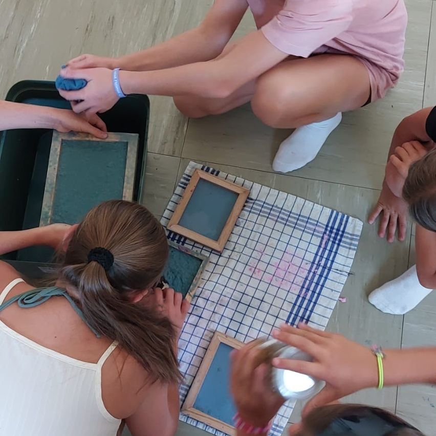 Eine Gruppe von Kindern sitzt auf dem Boden und ist in einer Kunstaktivität verwickelt. Sie verwenden eine Schale mit Ton und kleinen Holzrahmen. Ein Kind hält eine Schüssel mit Ton und ein anderes arbeitet an einem Tonstück.