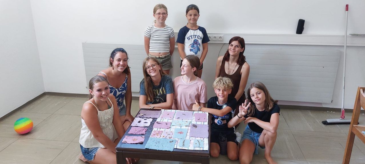 Eine Gruppe von Kindern und Erwachsenen lächelt und posiert für ein Foto um einen Tisch, der mit farbenfrohen Papierbasteleien bedeckt ist.