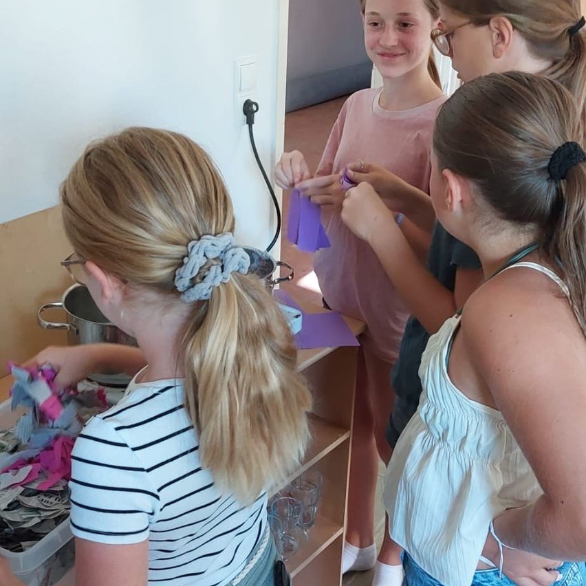 Vier Mädchen sind um einen Tisch versammelt und beschäftigen sich mit einer Bastelaktivität. Ein Mädchen hält ein lila Band, während ein anderes Mädchen eine Box mit Stoff hat. Der Raum hat eine weiße Wand und ein Holzregal.