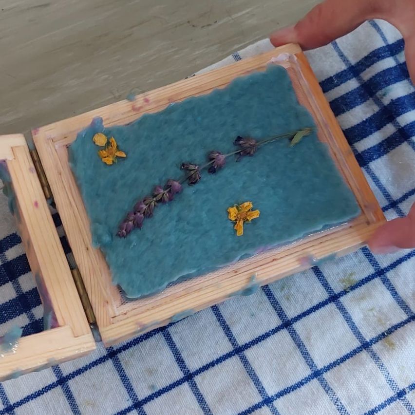 Eine Holzbox mit einer blauen Seifenbasis, verziert mit Lavendel- und gelben Blumen, wird von einer Hand auf einem blau-weiß karierten Tuch gehalten.