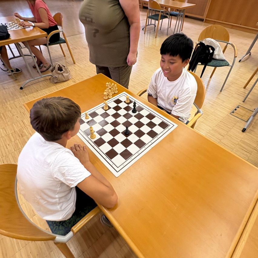 Zwei Jungen spielen Schach in einem Klassenzimmer. Ein Junge lächelt, während der andere sich auf das Spiel konzentriert. Eine Frau steht hinter ihnen.