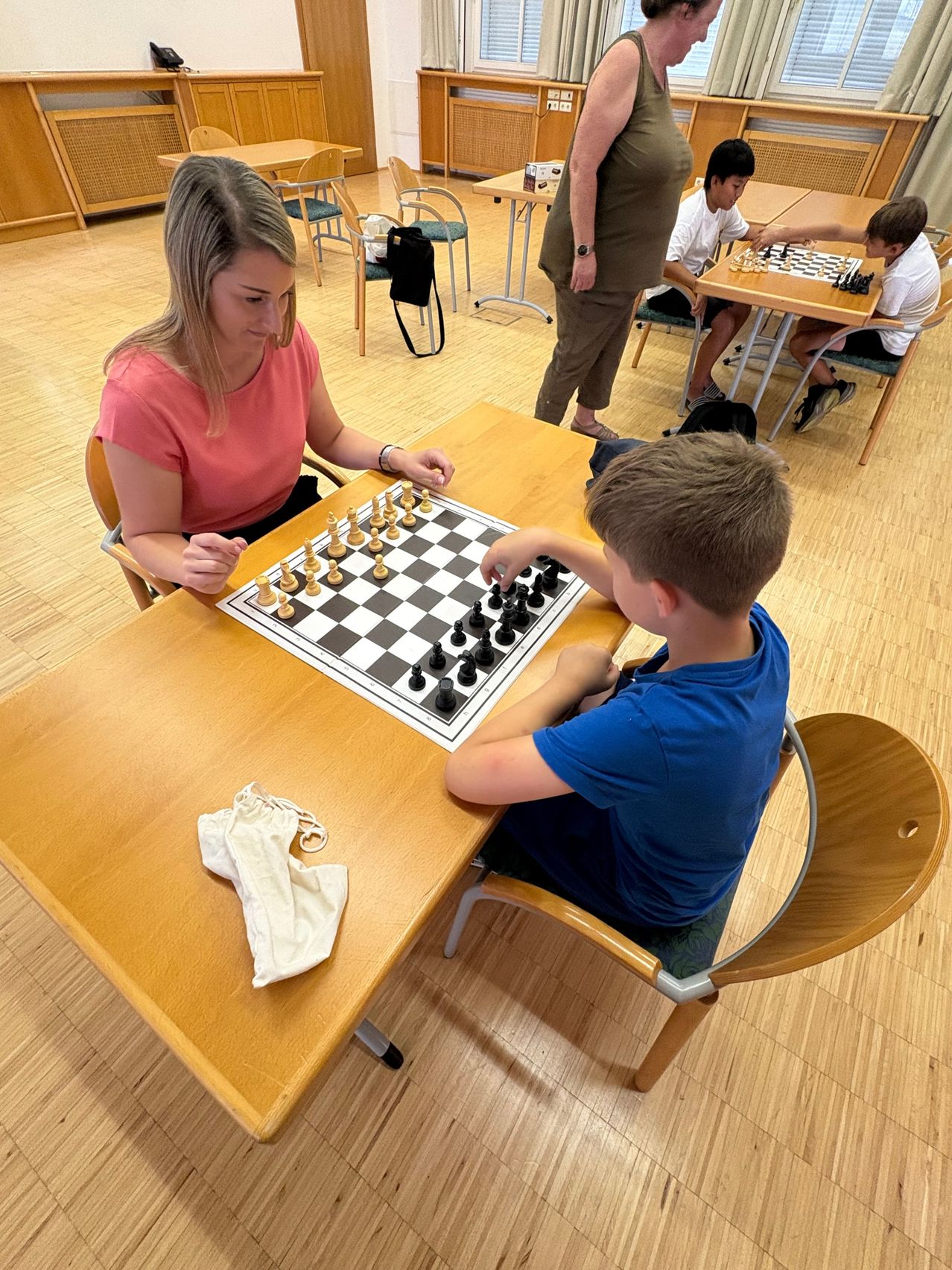 Eine Frau und ein junger Junge spielen Schach auf einem Holztisch in einem Raum, in dem andere Kinder Schach spielen.